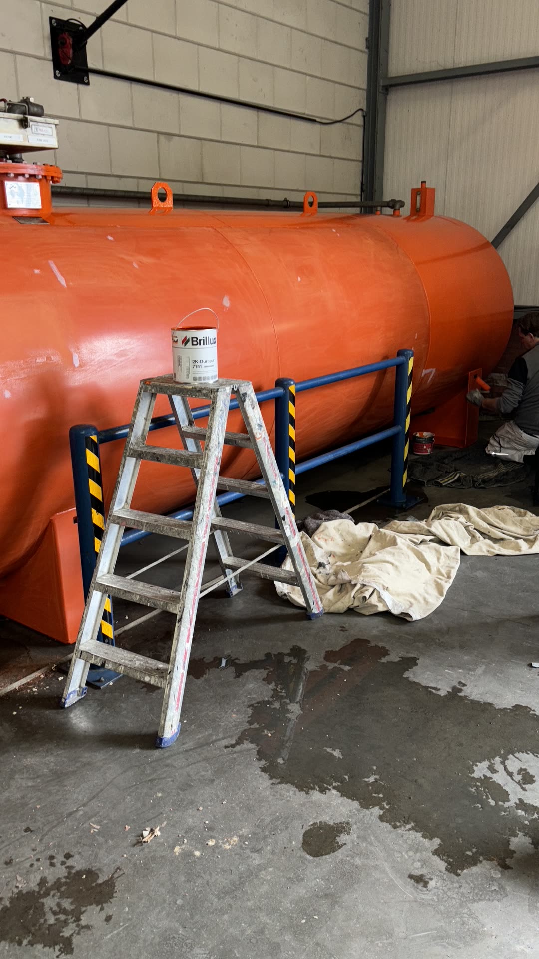 Oranje opslagtank in een bedrijfshal, met een trapje en een verfblik ernaast